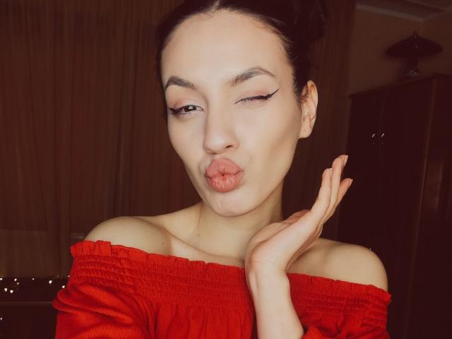 VievienneClemente with pouty lips, winking, and holding up her hand, wearing a red off-shoulder top, black hair in a bun, and bold eyeliner.