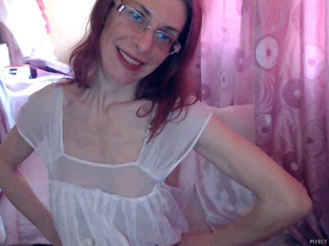 Une femme séduisante aux cheveux roux, portant des lunettes et une robe blanche transparente, souriante, montrant ses bras fins et sa petite poitrine, debout, les mains sur les hanches..