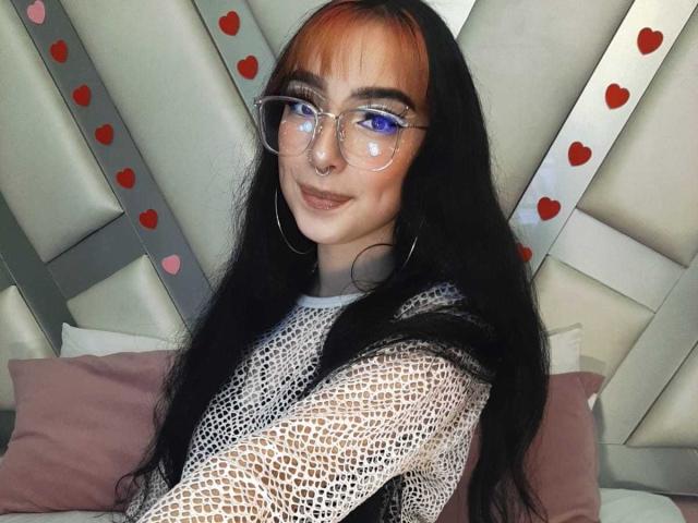 ConnieLuna, con lunghi capelli neri, frangia arancione, occhiali, piercing al naso, orecchini a cerchio e una maglietta bianca trasparente, sorride alla fotocamera, mostrando il suo viso carino e i suoi piccoli seni..