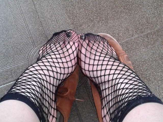 Os pés de SammyFour, com meias de rede preta, com os dedos levemente curvados, usando botas marrons de cano curto, em um chão cinza texturizado. Unhas dos pés visíveis, pele clara e um padrão de rede solto..