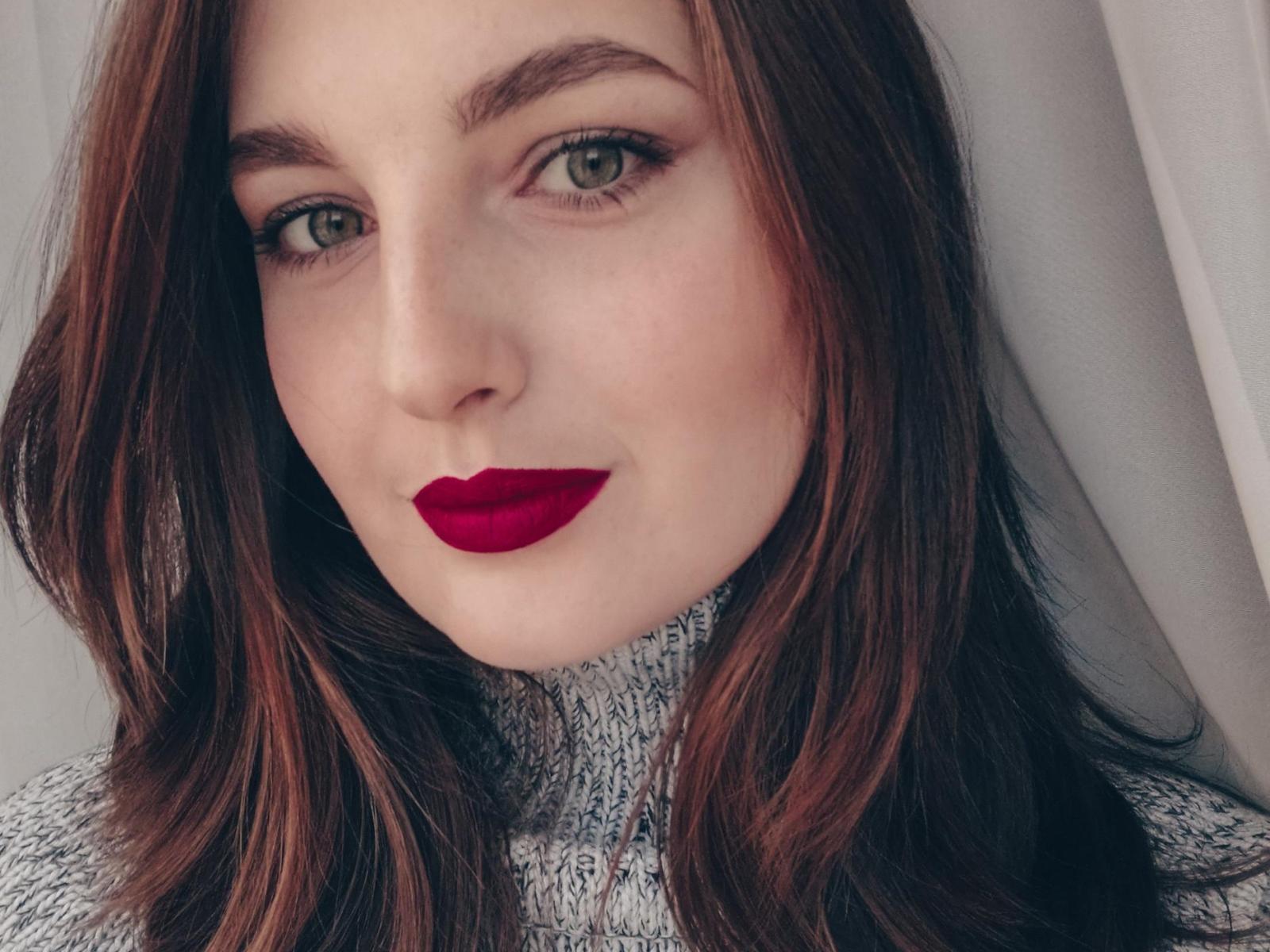 Outono. O rosto de Sara, com batom vermelho, olhos verdes e cabelo castanho ondulado, vestindo um suéter cinza de gola alta. Ela está olhando para a câmera com um sorriso leve e sedutor..