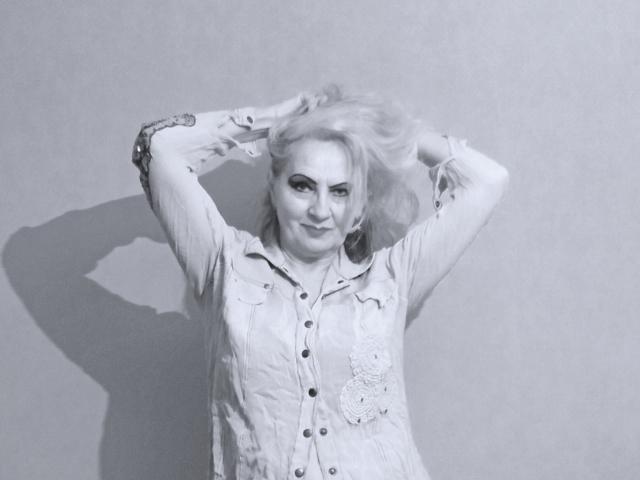 CarlyTreat hat blondes Haar, trägt ein geblümtes Hemd, das zugeknöpft ist, hat die Hände im Haar, lächelt, grauer Hintergrund, Schatten an der Wand. Ihre Brüste sind klein, sie ist süß..