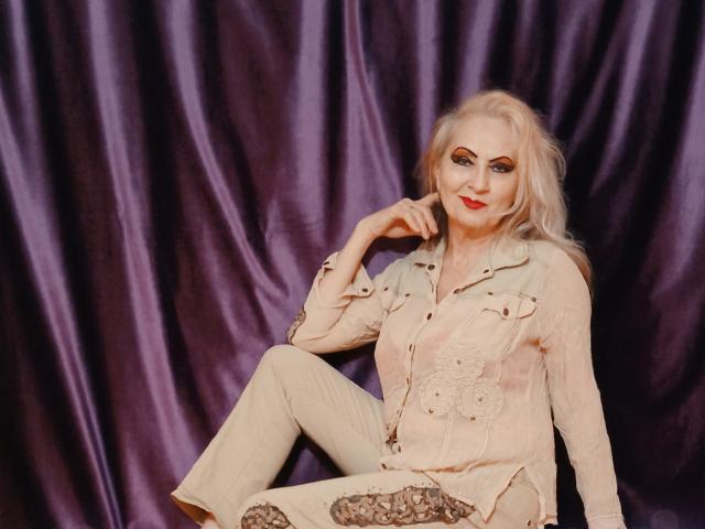 Carly, avec ses cheveux blonds, son rouge à lèvres rouge et sa tenue beige, assise devant des rideaux violets, une main sur sa joue, souriante, portant une chemise boutonnée et un pantalon, montrant subtilement sa poitrine..
