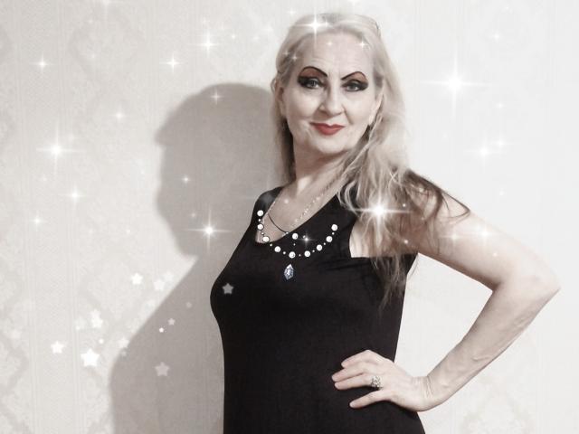 Carly, con lunghi capelli biondi, vestito nero senza maniche, collana con una pietra blu, sorridente, con una mano appoggiata al fianco, effetti di luce scintillante, sfondo bianco. Il seno è leggermente visibile sotto il vestito..