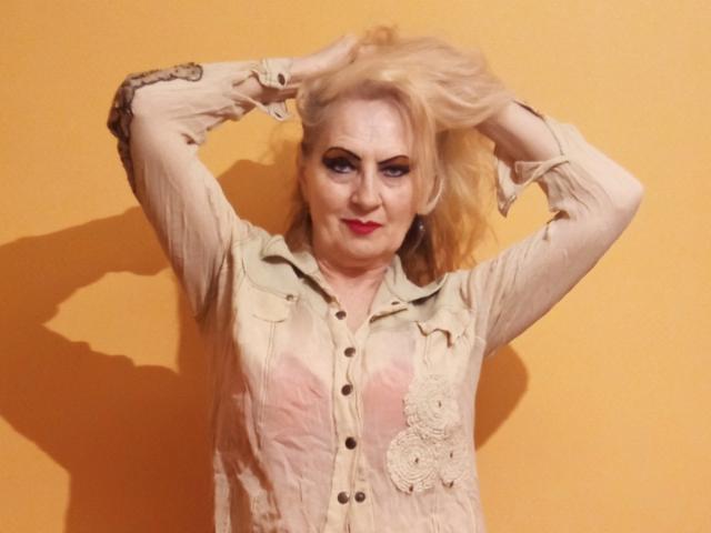 CarlyTreat tem cabelo loiro, olhos azuis, batom vermelho e está vestindo uma camisa bege com botões, com as mãos no cabelo, em pé em frente a uma parede amarela, com uma expressão confiante..