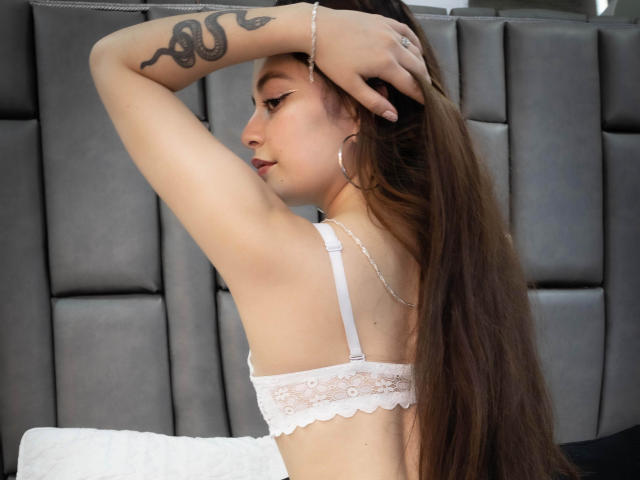 DaniaMiller with long brown hair, white lace bra, and hoop earrings, arms raised, showing snake tattoo, looking side profile, silver chain on right arm.