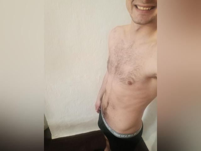 AlexAhSunny, sem camisa, com o peito peludo, sorrindo, puxando a calça preta para baixo para mostrar um pouco de seu pênis flácido e circuncidado, em pé contra uma parede branca..