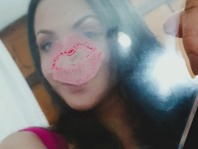 ElinaJhonson's face with pink lipstick kiss mark on a glass, holding a phone in a pink top, black hair, and a slightly blurred background.