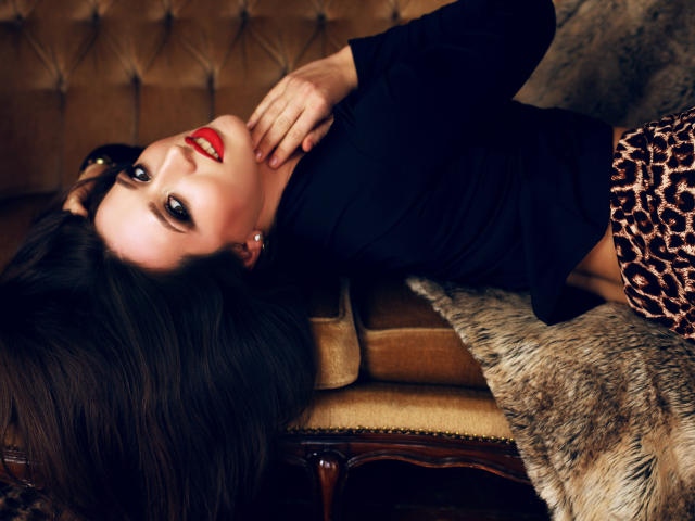 Bonney with red lipstick, lying on a brown tufted sofa, black top, leopard print skirt, hand on chest, dark hair, looking up, fur blanket, sexy, dark eyes, soft lighting.