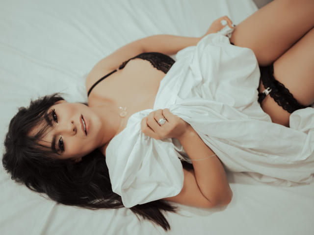 Briana Preston, con un sujetador y tanga de encaje negro, acostada en una cama blanca, sosteniendo una sábana blanca, mostrando sus pechos y vulva, cabello negro largo, un anillo en su mano izquierda, mirando a la cámara..