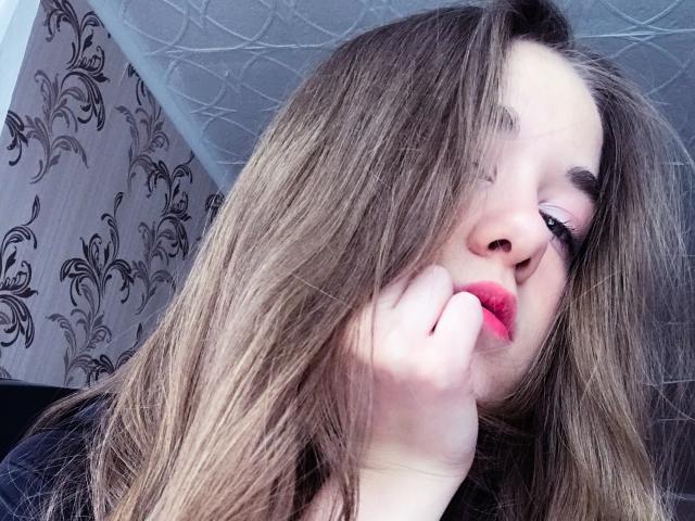 ToryMolly tiene el cabello castaño cubriendo un ojo, lápiz labial rojo y está tocándose la cara con una mano. Tiene pestañas largas y una expresión ligeramente sensual y con los labios fruncidos..