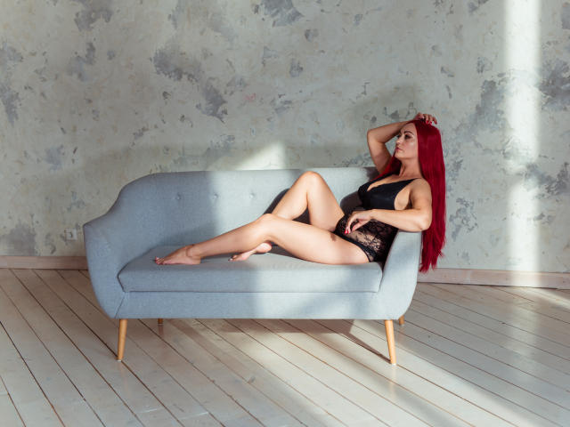 VioletNice with long red hair, black lace lingerie, and bare feet, lounging on a blue couch, one arm raised, legs crossed, showing her ass and tits.