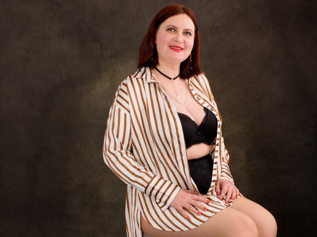 BigTitsXHot, avec des cheveux roux, souriante, portant une chemise rayée ouverte pour montrer un soutien-gorge et des sous-vêtements noirs, assise, les mains sur les genoux, avec un collier, sur un fond sombre..