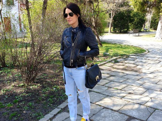 JoliDanielle, con i capelli scuri, indossa occhiali da sole, una giacca di pelle nera, jeans azzurri strappati, una cintura nera, una borsa nera, è in piedi su un sentiero di pietra, con le mani nelle tasche, e guarda in avanti..
