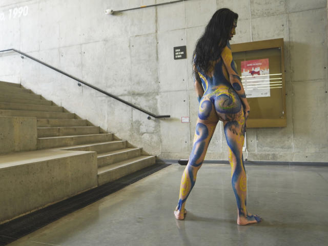 BellaMariee, naakt, met blauwe en gele bodypaint, staat in een betonnen gang en toont haar beschilderde achterwerk en lange donkere haar, terwijl ze wegkijkt van de camera..