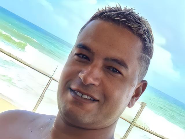MrBlackHot, senza camicia, con i capelli castani corti, sorride alla telecamera, ha un viso paffuto e la pelle abbronzata, ed è in piedi vicino all'oceano, con acque blu cristalline sullo sfondo..