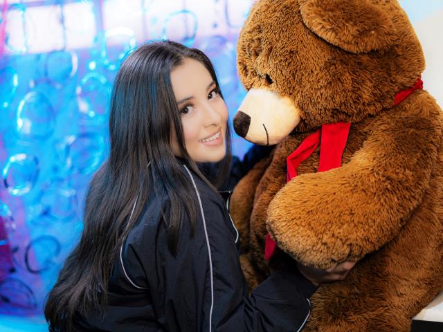 SantaRodrigues mit langem, schwarzem Haar, lächelnd, hält einen riesigen, braunen Teddybären mit einem roten Band, trägt eine schwarze Jacke mit weißer Verzierung. Brüste, Hintern, Brüste sind nicht sichtbar..