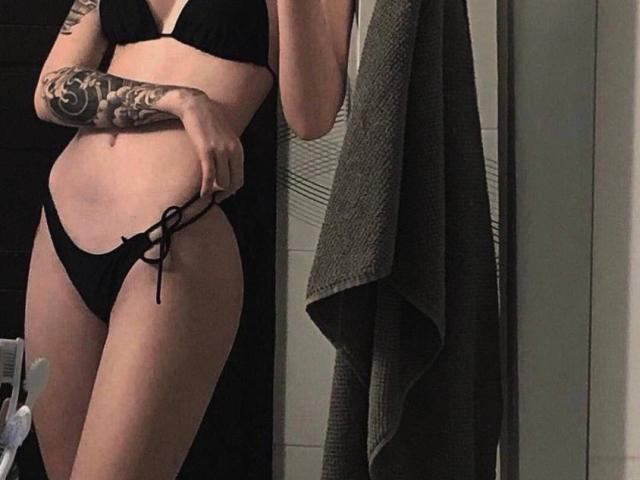 Diana, usando um biquíni preto, mostrando seu traseiro e seios pequenos, tem um braço tatuado e está segurando uma toalha em um banheiro com pouca luz. Sua mão está em sua cintura..