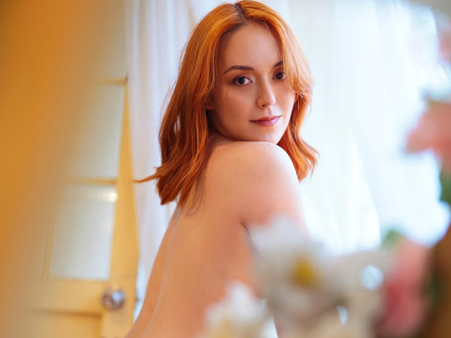 Molly Marmalade mit roten Haaren, die über ihre Schulter schaut, nackt, mit einem sanften Lächeln, heller Haut und kleinen Brüsten, in einem hellen Raum, verschwommene Blumen im Vordergrund..