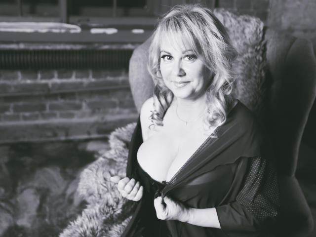 Black-and-white photo of BeverlyMiller with curly blonde hair, large tits, and a confident smile, unzipping a dark dress, exposing black lace lingerie, seated in a furry chair.