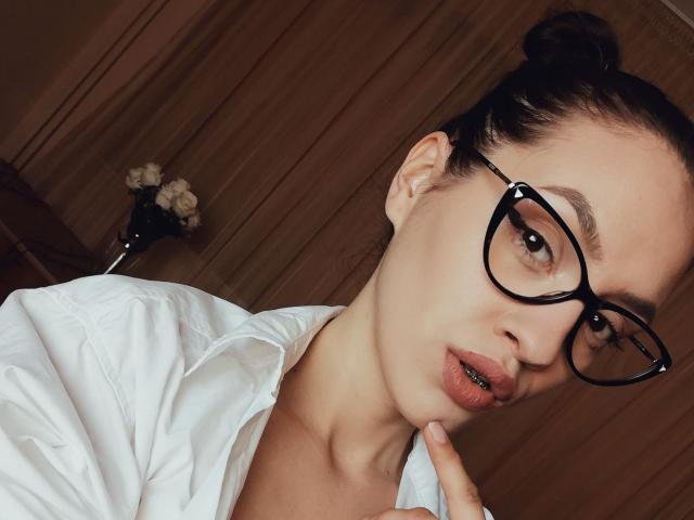 VievienneClemente with black glasses, white shirt, and her finger on her chin, showing her pouty lips and updo, has a sexy, nerdy look with her ass in the background.