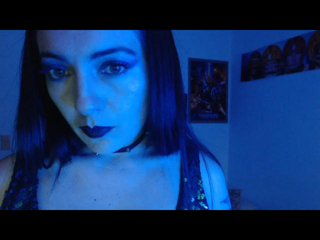 PollyAnnaX con il trucco blu scuro, il rossetto nero, una maglietta con paillettes e una collana nera, guarda la fotocamera in una stanza illuminata di blu. Il suo seno è leggermente visibile, con una luce blu..