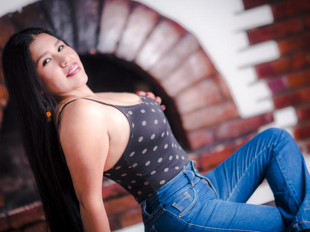 AgathaPember with long black hair, wearing a polka dot tank top and blue jeans, sitting with her back slightly arched, showing her tits, smiling at the camera.