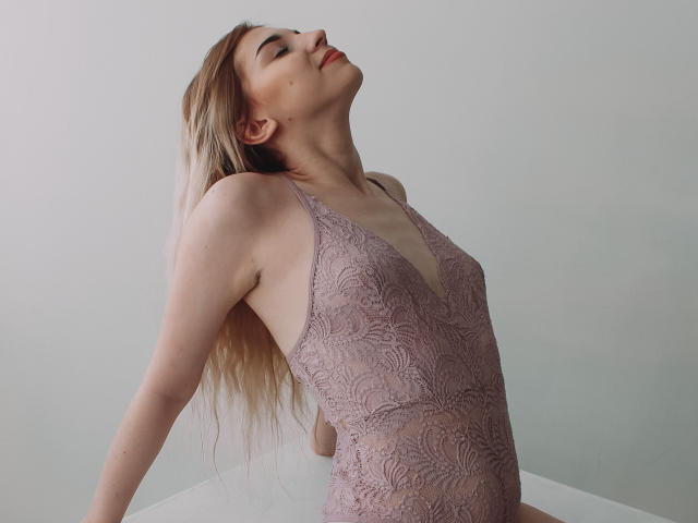 ScarletAmi, blond, mit geschlossenen Augen und geneigtem Kopf, trägt einen pinken Spitzen-Body, der ihre kleinen Brüste und ihren schlanken Hintern zeigt, und lehnt sich an eine weiße Oberfläche, wobei ihre Arme hinter ihr sind..