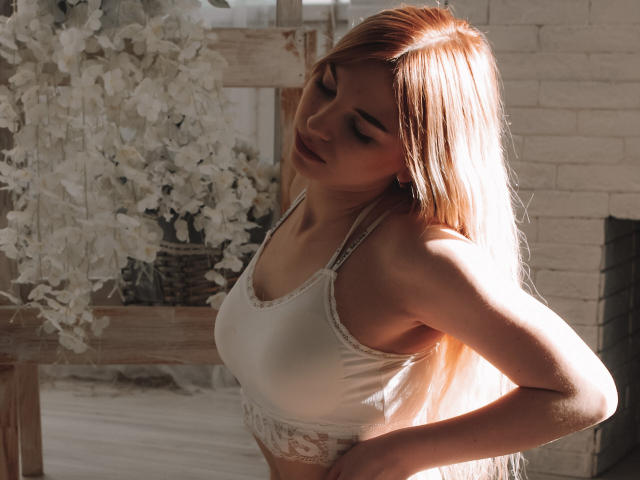 ScarletAmi, con cabello rubio largo, ojos cerrados, vistiendo un sostén de encaje blanco, la luz del sol proyectando suaves sombras sobre ella, sus pechos ligeramente visibles, sus glúteos no son visibles..