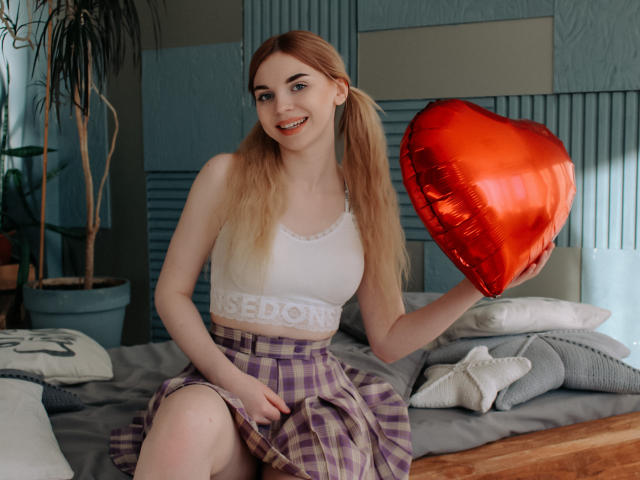 ScarletAmi with blonde pigtails, white crop top, purple plaid skirt, holding red heart balloon, smiling, sitting on bed with pillows, looking cute, showing small tits.