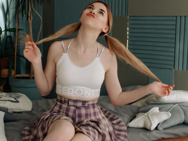 ScarletAmi con coletas, sostén blanco con encaje, y falda de cuadros, sentada en una cama, sujetándose las coletas, sonriendo, piel pálida, cabello rojo, pechos pequeños, linda, dormitorio..
