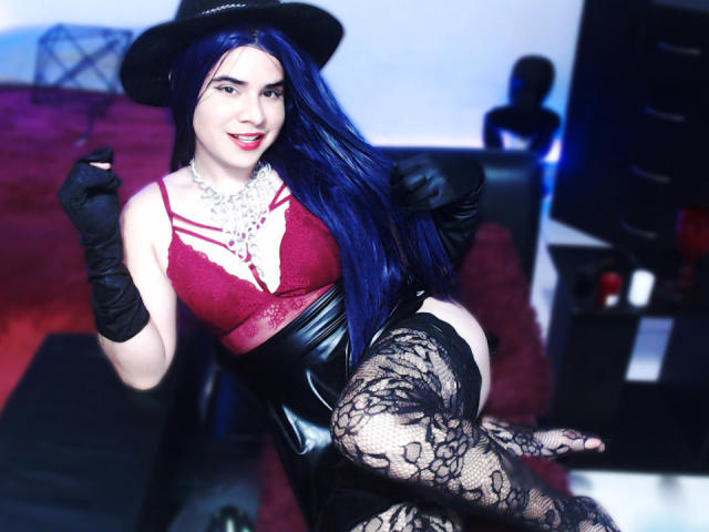 CamilaFetishDirty with blue hair, red lace bra, black leather corset, lace stockings, black gloves, and hat, smiling, showing tits, sitting, holding glass, in sexy, seductive pose.