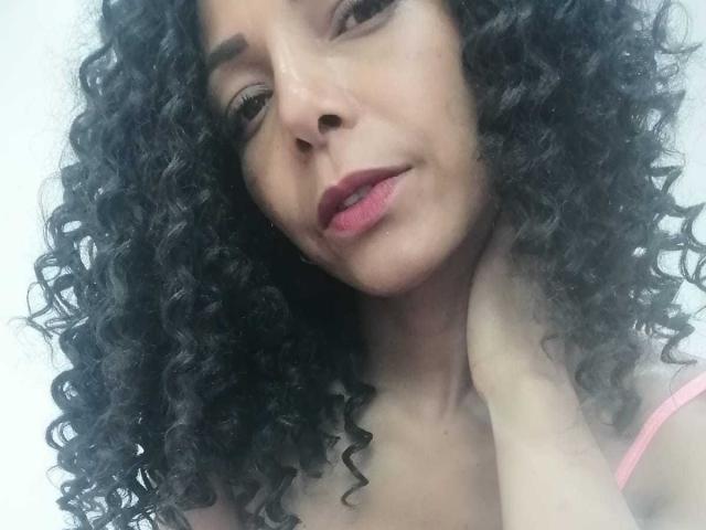 VenusChaud, com cabelos pretos cacheados, batom rosa e um leve sorriso, mostrando seu pescoço e a alça rosa. Suas mãos estão perto da garganta. Ela está olhando para baixo..