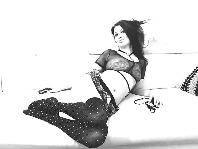 JoliDanielle is lying on a white couch, wearing a sheer top, black panties, and polka-dot tights, with her ass up and legs crossed, holding a pair of glasses.