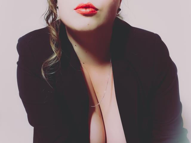 JuliaFrancaise con rossetto rosso, un blazer nero e una camicia scollata che mostra il décolleté, una sottile collana e i suoi capelli castani ondulati che le ricadono sulla spalla sinistra..
