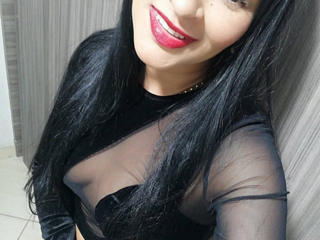 AlyssiaOcean with black sheer top, visible black bra, red lipstick, and long black hair, smiling. Boobs slightly shown through sheer top. Background is grey wood panel.
