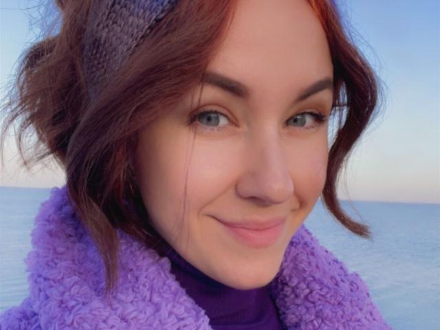 Misstyk has light skin, blue eyes, and reddish-brown wavy hair, wearing a purple fuzzy scarf and headband, smiling subtly with a cute nose and a slightly curved lip.