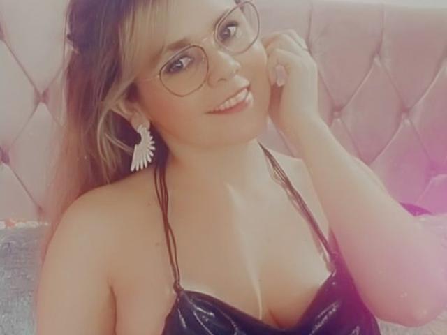 Milek with glasses, black vinyl top, showing her tits, smiling, hand on cheek, pink tufted headboard, feather earrings, blonde hair, pinkish light, soft focus, cute.