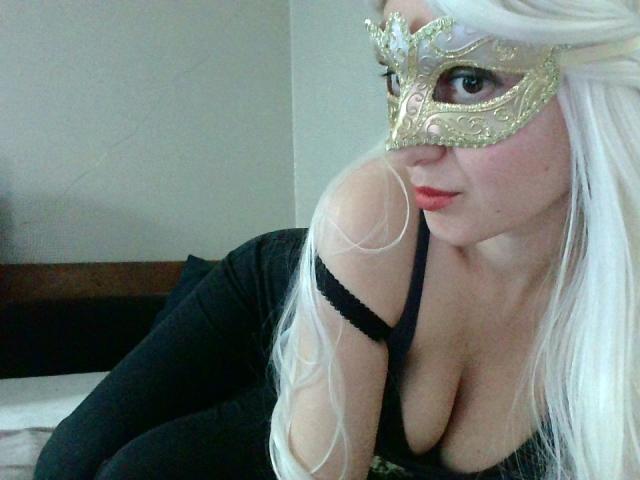 SophieLajoie met lang wit haar, die een gouden masker draagt, een zwarte top met open schouders en zwarte broek, waarbij haar grote borsten zichtbaar zijn, leunt voorover op een bed..