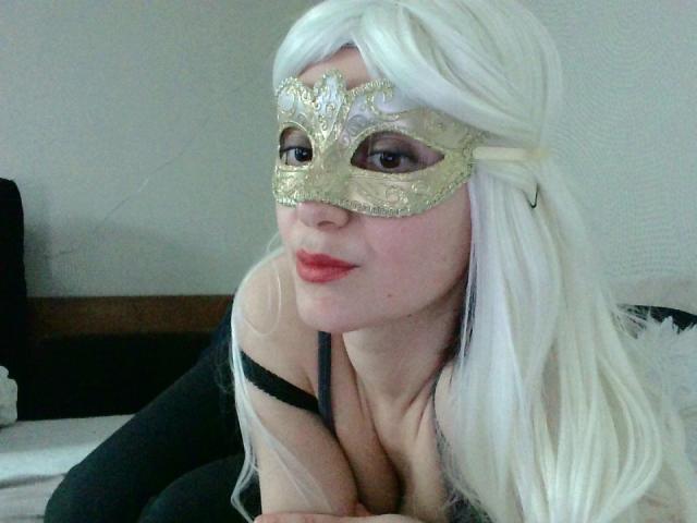 SophieLajoie met lang wit haar, een gouden masker, een zwarte top en rode lippen, leunt voorover en laat haar borsten zien, op een bed..