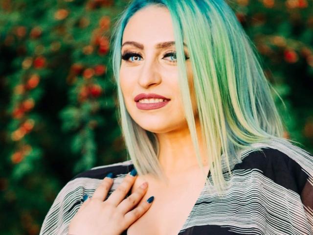 Agaa con capelli verde-blu, camicia a righe, unghie blu, che sorride, con una mano sul petto, che mostra leggermente il seno, rossetto rosa, occhi azzurri, che guarda di lato, sfondo esterno..