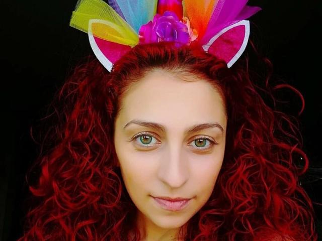 Agaa, met krullend rood haar, draagt kleurrijke, regenboogkleurige kattenoren en een paarse roos. Ze heeft groene ogen en een subtiele glimlach en kijkt recht in de camera..