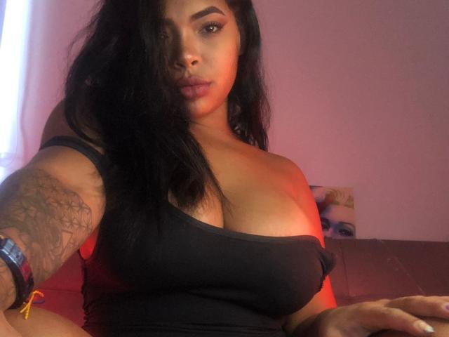 VickyGreys with black hair, tan skin, and large tits, wearing a black tank top, tattoo on arm, red light, and a serious expression, sitting on a brown sofa.
