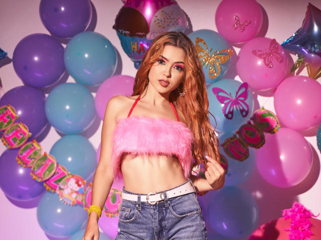 MaisyWarner with long pinkish-red hair, wearing a fluffy pink strapless top, exposing her small tits, and high-waisted blue jeans, standing in front of colorful balloons.