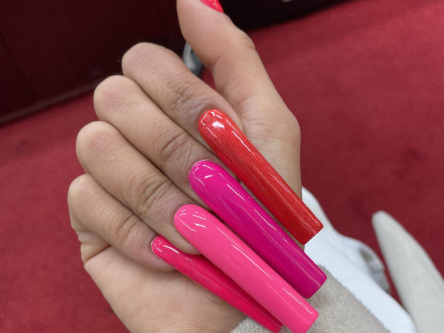 MissFeetParis' hands with long, glossy, pink and red nails, stacked and glossy, fingers slightly curled, holding her hand up. Red carpet background.