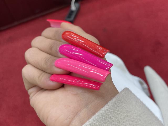 La main de MissFeetParis, ornée d'ongles acryliques longs, roses et rouges, empilés les uns sur les autres, exhibe son vernis à ongles brillant, rose vif et rouge, sur un tapis rouge..