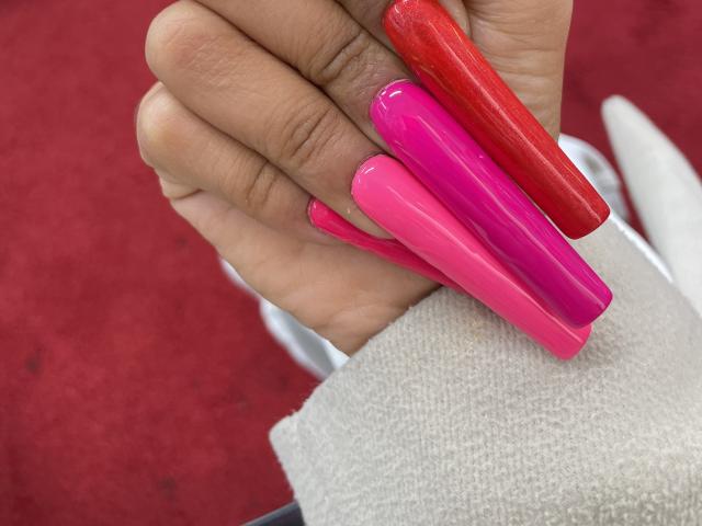 A mão de MissFeetParis segura duas unhas postiças longas e brilhantes, em vermelho e rosa. Suas unhas estão sobre uma toalha branca, com um tapete vermelho ao fundo..