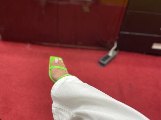 I pantaloni bianchi di MissFeetParis e i tacchi a punta verde brillante con lo smalto bianco sui piedi, sul tappeto rosso, che mostra la pianta del suo piede in una posa rilassata e leggermente inclinata..