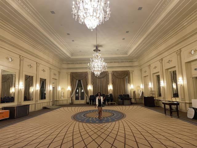 Uma mulher de pele morena, cabelos pretos e seios médios está em um quarto luxuoso, com dois lustres de cristal, paredes bege e um tapete estampado..