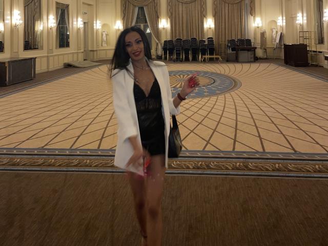 MissFeetParis met lang zwart haar, gekleed in een witte jas over een zwarte lingeriejurk, houdt een rode appel vast en staat in een grote kamer met tapijten op de vloer..